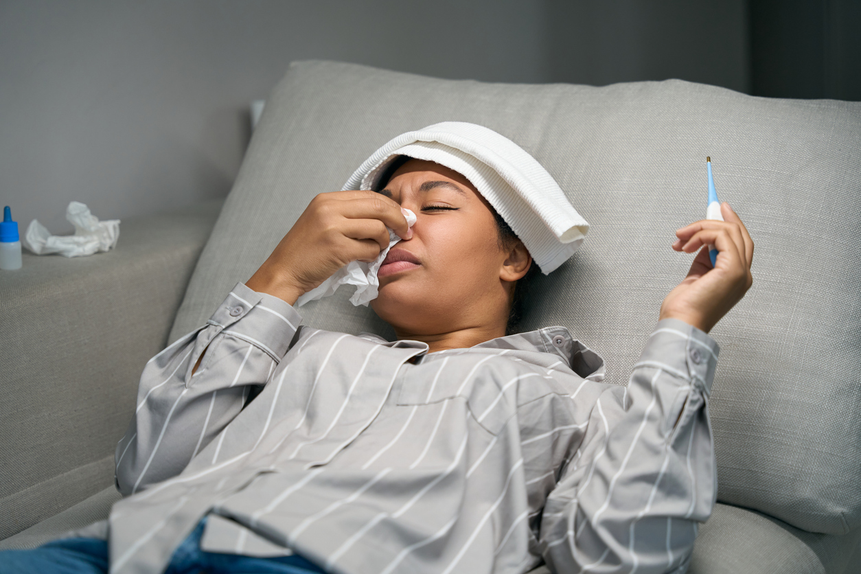 Woman resting on her couch at home with cold and flu symptoms, holding a thermometer